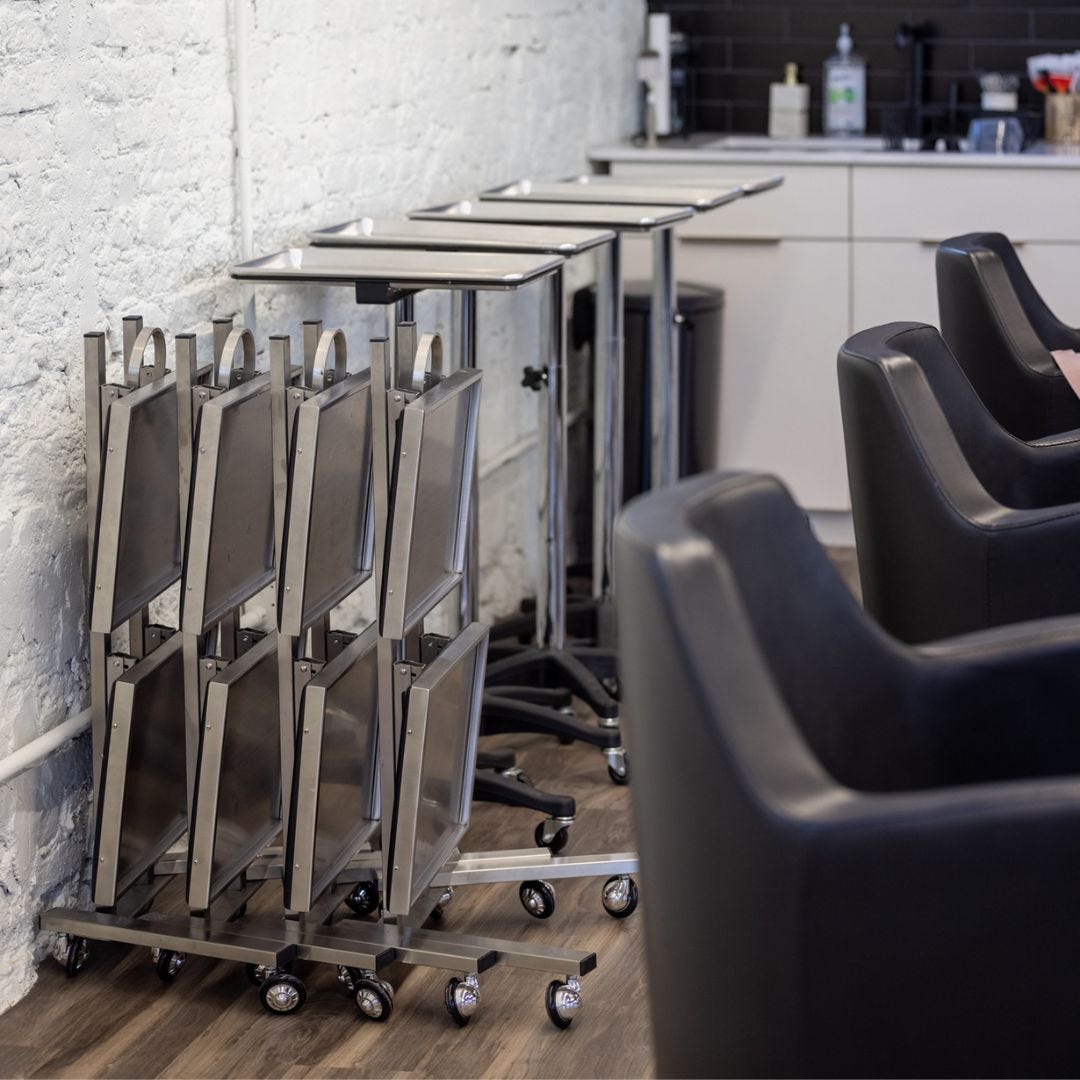 four adjustable-height aluminum hair salon service trays along a wall and four collapsible service trays with 2 trays each lined up along a wall behind styling chairs