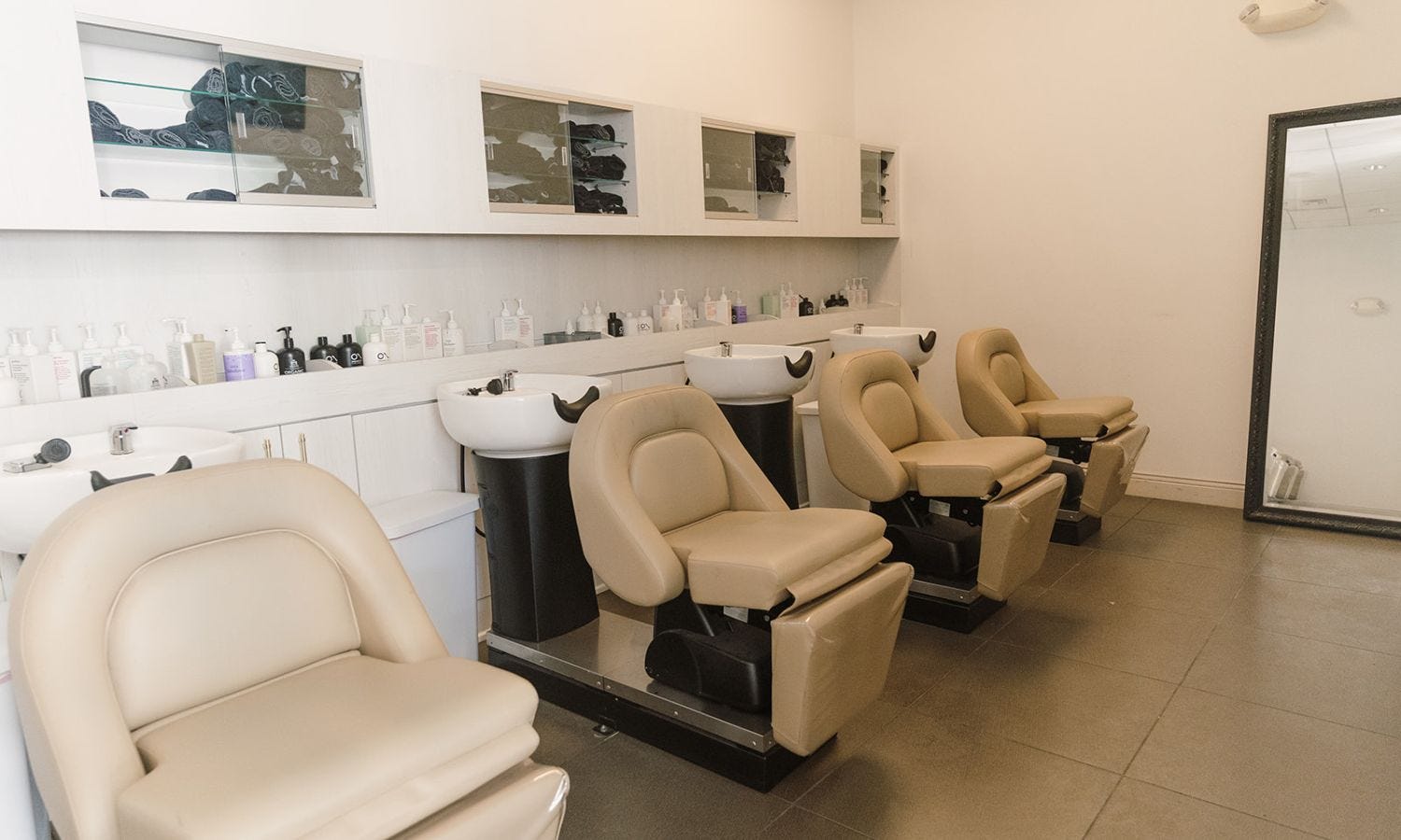 four electric shampoo sidewash units with cabinets above full of towels and a large mirror at the end of the room